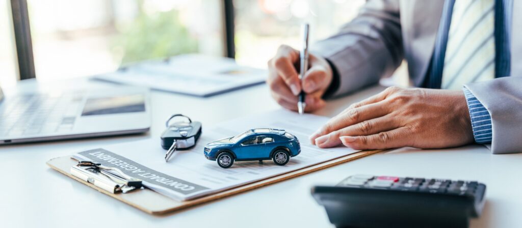 Person signing a car finance agreement with a miniature car model and keys on a desk, symbolizing the process of securing auto loans.