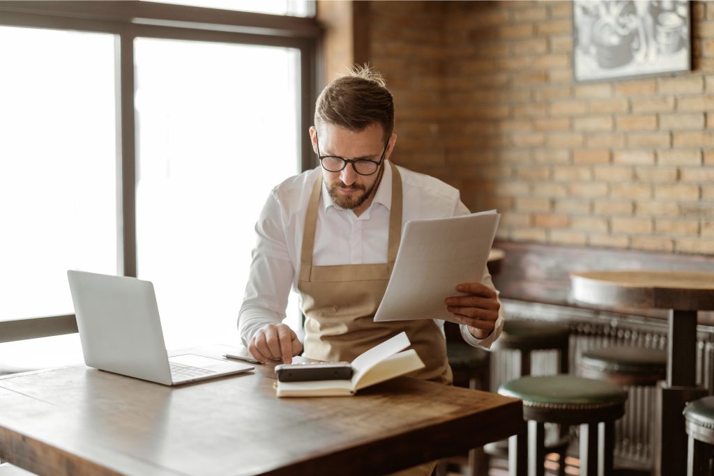 Man in an apron reviewing financial documents at a wooden table with a laptop and notebook, illustrating the process of managing home loans and financing options in a café setting.