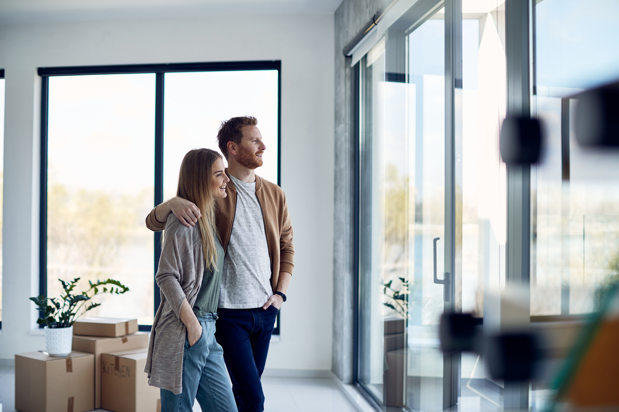 Couple smiling and embracing in a bright, modern room with large windows, surrounded by moving boxes, symbolizing first home ownership in Sydney.