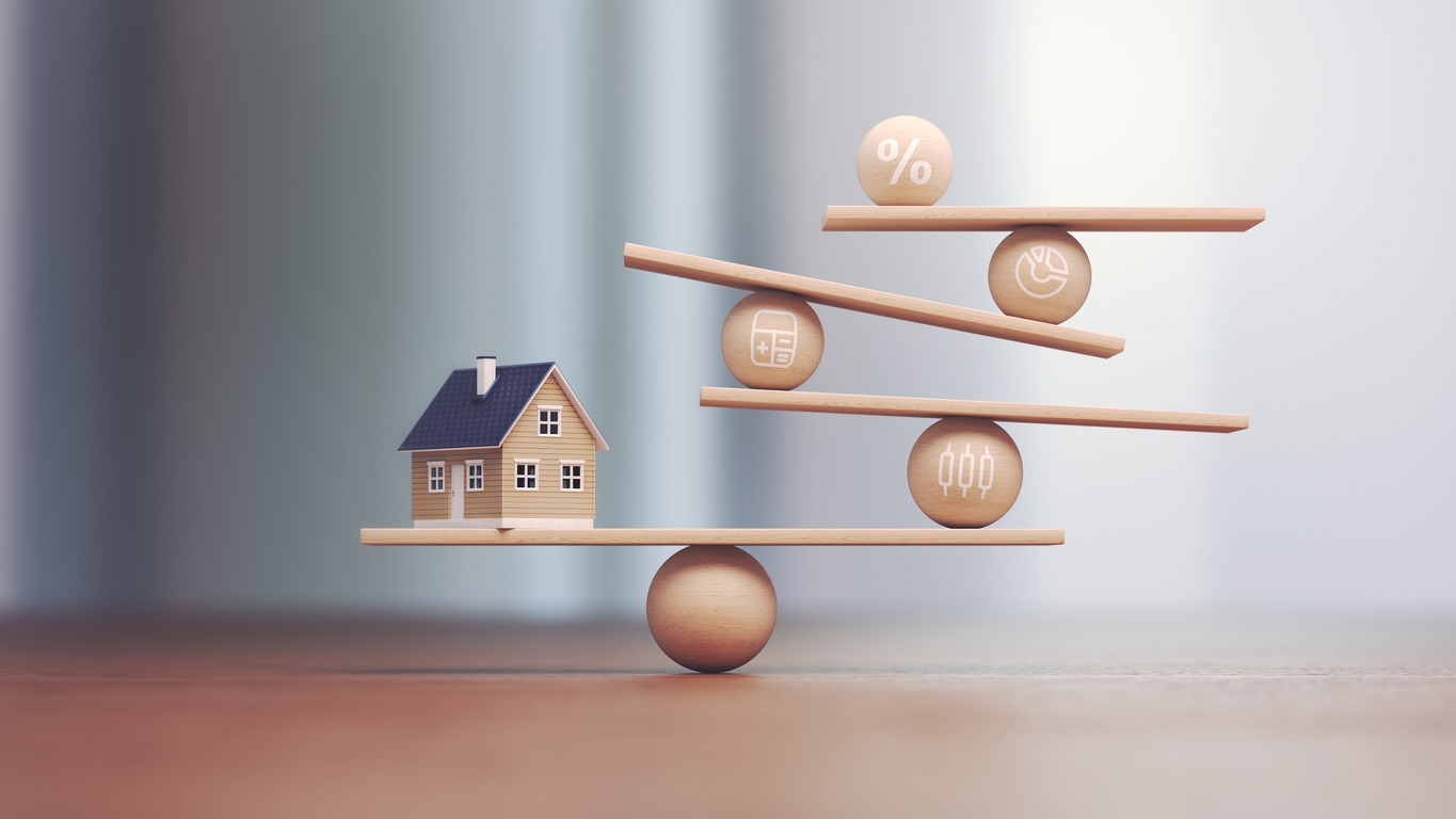 Wooden balance scale with a miniature house, percentage symbol, calculator, and financial icons, representing the comparison of fixed and variable rate business loans for construction projects.