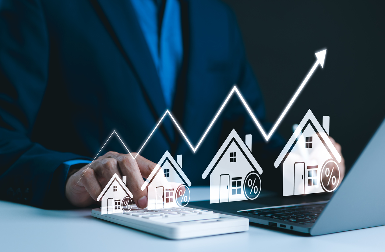 Business professional analyzing investment property loan trends with a calculator and laptop, featuring graphical representations of houses and upward financial growth indicators.