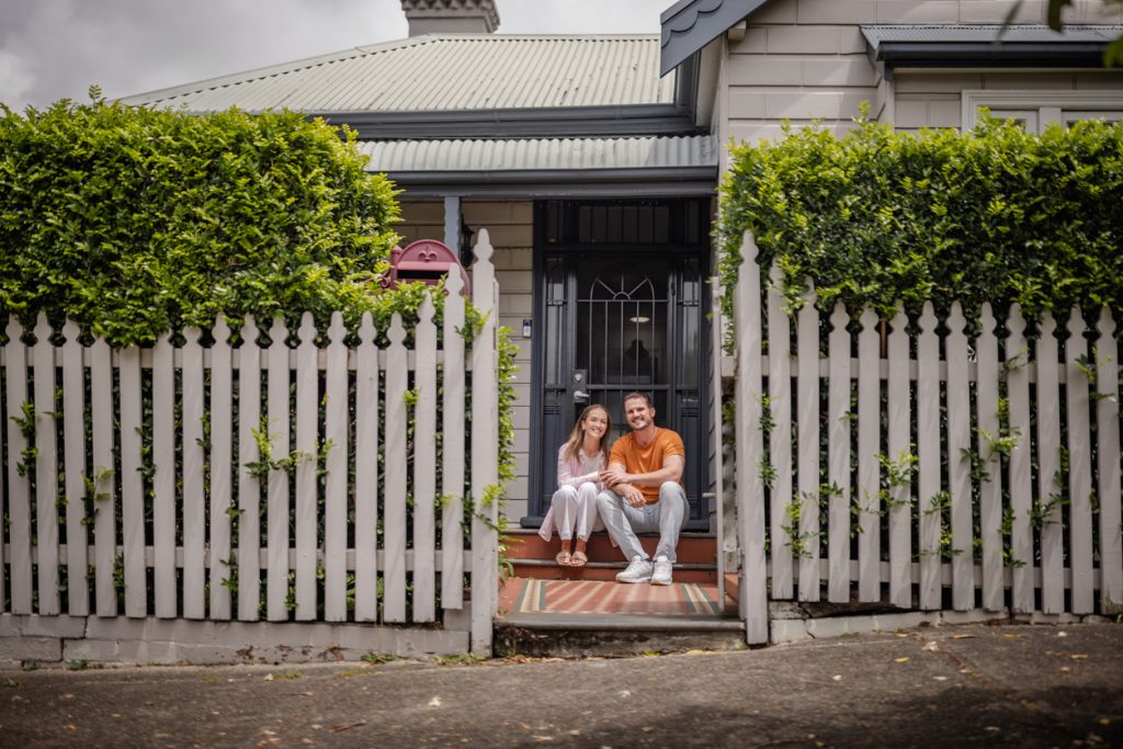 Couple sitting on the steps of a house, surrounded by a white picket fence and lush greenery, symbolising home ownership and financial stability in the context of home loans.