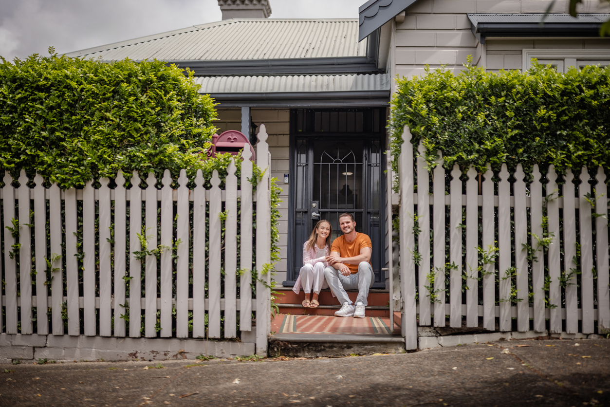 Couple sitting on the steps of a charming home in Sydney, surrounded by a white picket fence and lush greenery, representing first home buyer aspirations.