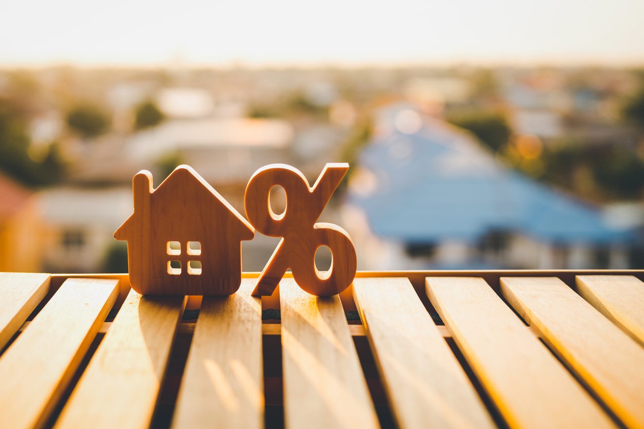 Wooden house and percentage symbol on a table, symbolizing interest rates related to home loans, against a blurred urban background.