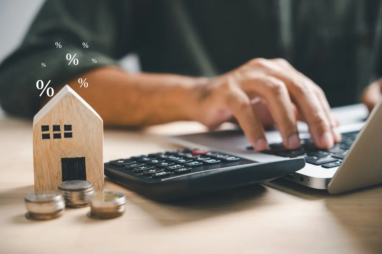 Person calculating refinancing options on a laptop, with a wooden house model and coins, representing home loan refinancing and financial planning.