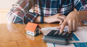 Person using a calculator with mortgage documents and a small model house, illustrating financial calculations related to home refinancing.