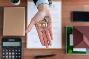 Hand holding house keys above a contract, calculator, pen, and a model house, symbolising home loan processes and direct bank services.