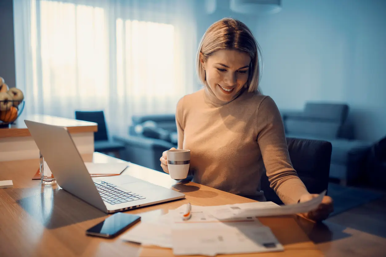 Woman reviewing documents while sitting at a desk with a laptop and a coffee cup, representing home loan refinancing and financial planning in a cozy setting.