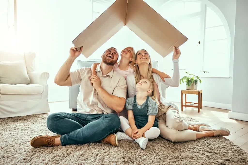 Family celebrating in a living room, holding a cardboard roof, symbolising home ownership and financial security related to mortgage refinancing.