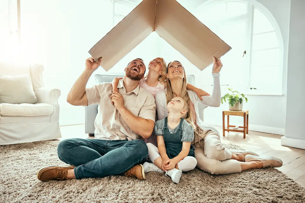 Family smiling while holding a cardboard roof in a cozy living room, symbolizing homeownership and family togetherness, relevant to investment property refinancing.