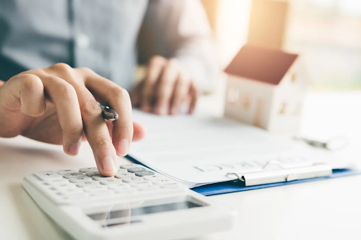 Person using a calculator while reviewing mortgage documents and a model house, illustrating financial planning for home loans and refinancing options.