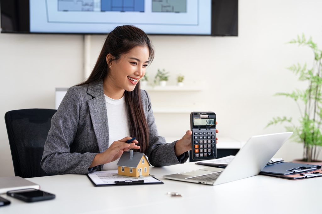 Woman in a grey blazer using a calculator and holding a model house, with a laptop and documents on a desk, in an office setting related to home loans and refinancing options in Sydney.