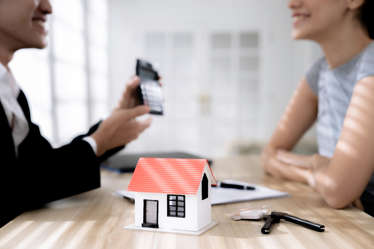 Man discussing SMSF home loans with a woman, calculator in hand, miniature house model on table, documents and keys nearby, illustrating property investment options.